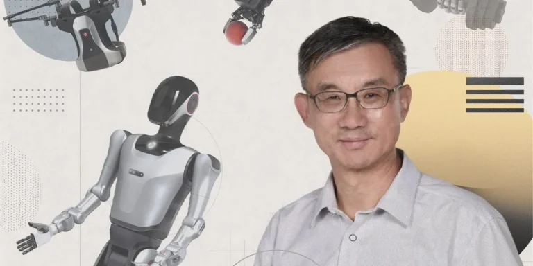 man wearing glasses and a gray shirt smiles at camera while surrounded by futuristic robots and tech